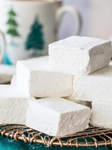 square-shaped homemade marshmallows stacked on a gold metal cooling rack with mugs of hot chocolate in the background