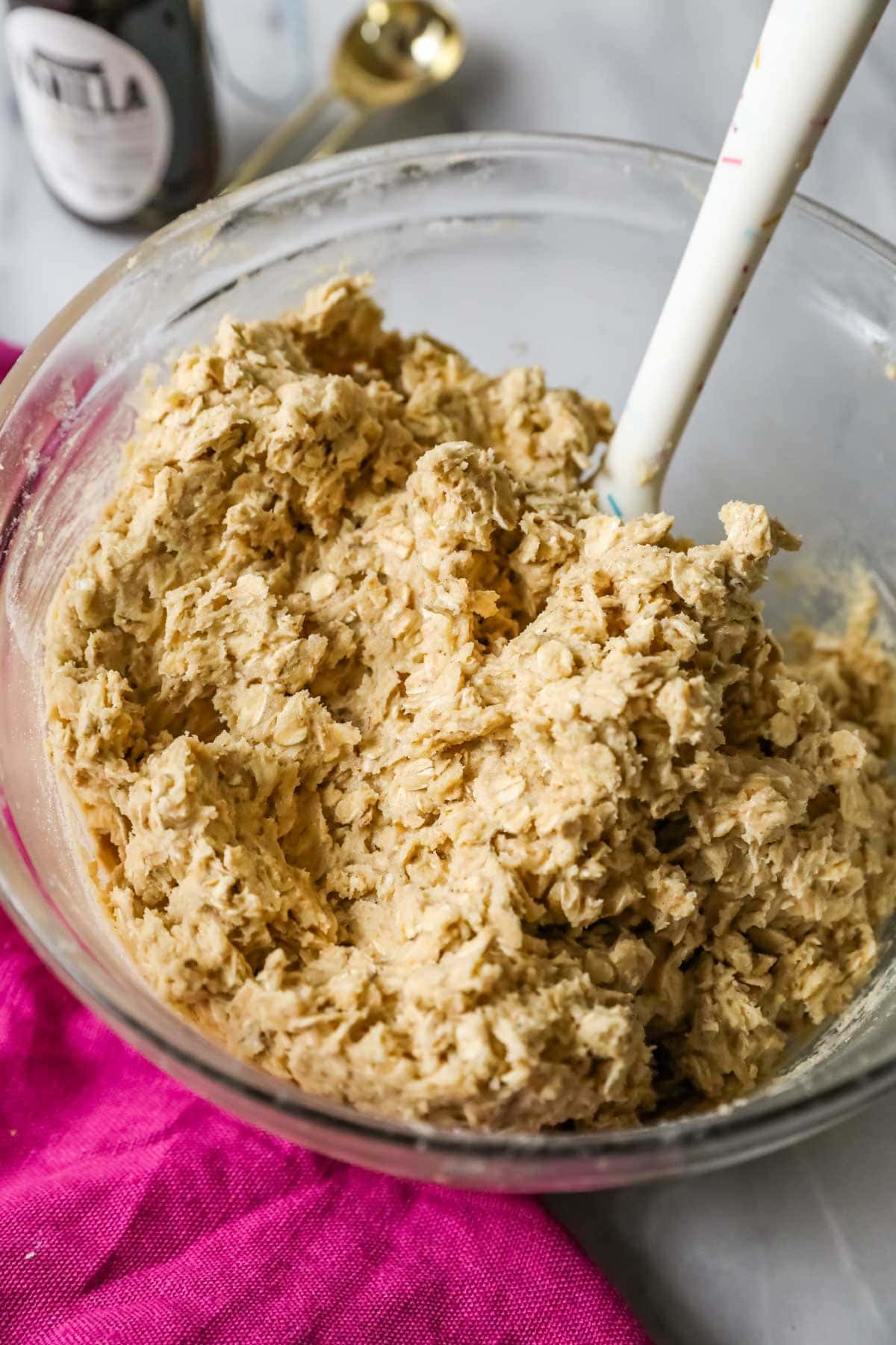 Oatmeal cookie dough in a glass bowl, pink towel in foreground, ingredients in background