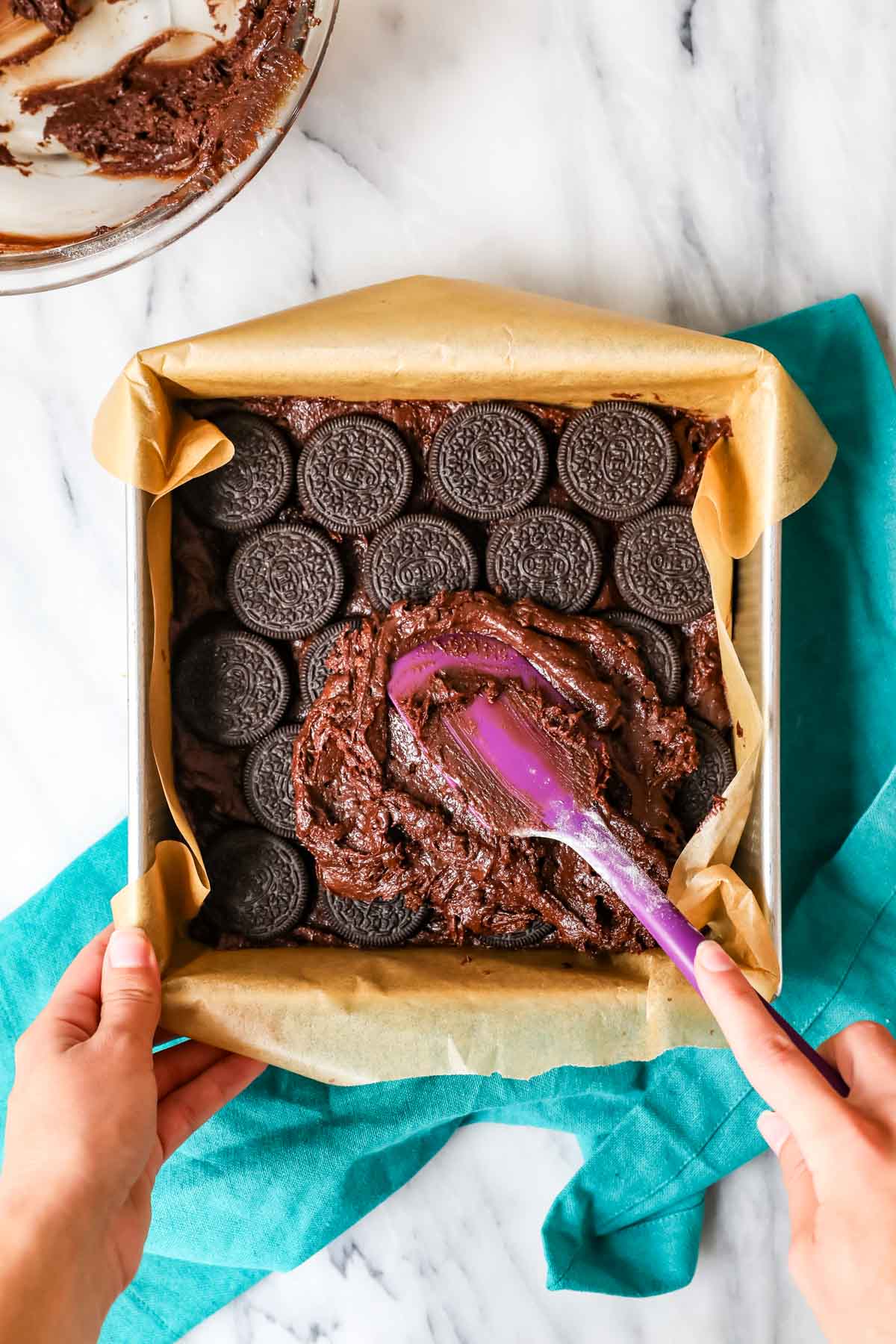 Overhead view of brownie batter being spread over Oreos in a square parchment lined pan.