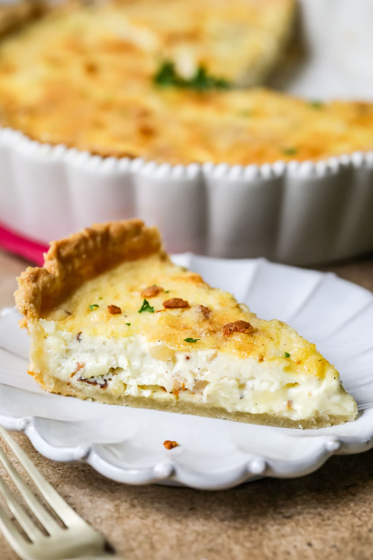 Slice of quiche on a white plate with the remaining quiche behind it.