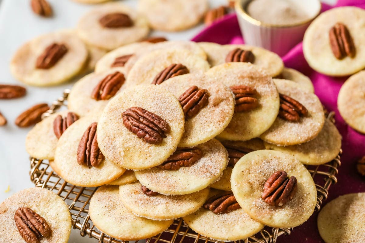 Thin cookies dusted with cinnamon sugar and topped with pecan halves stacked in a pile on a cooling rack.
